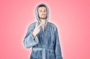 Portrait of young caucasian bearded man in blue bathrobe shows victory or peace gesture isolated on pink background photo