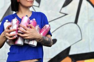 Portrait of an emotional young girl with black hair and piercings. Photo of a girl with aerosol paint cans in hands on a graffiti wall background. The concept of street art and use of aerosol paints