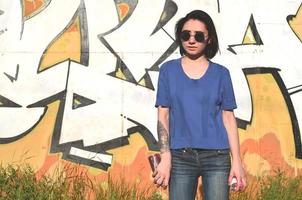 retrato de una joven emocional con pelo negro y piercings. foto de una chica con latas de pintura en aerosol en las manos sobre un fondo de pared de graffiti. el concepto de arte callejero y uso de pinturas en aerosol