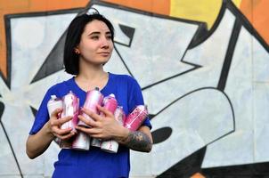 Portrait of an emotional young girl with black hair and piercings. Photo of a girl with aerosol paint cans in hands on a graffiti wall background. The concept of street art and use of aerosol paints