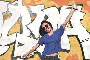 Portrait of an emotional young girl with black hair and piercings. Photo of a girl with aerosol paint cans in hands on a graffiti wall background. The concept of street art and use of aerosol paints