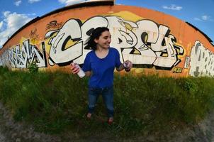 Portrait of an emotional young girl with black hair and piercings. A wide-angle photo of a girl with aerosol paint cans in the hands on a graffiti wall background. A modern portrait of a fisheye lens