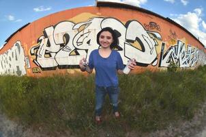 Portrait of an emotional young girl with black hair and piercings. A wide-angle photo of a girl with aerosol paint cans in the hands on a graffiti wall background. A modern portrait of a fisheye lens