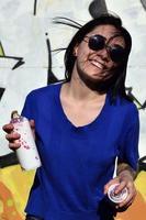 retrato de una joven emocional con pelo negro y piercings. foto de una chica con latas de pintura en aerosol en las manos sobre un fondo de pared de graffiti. el concepto de arte callejero y uso de pinturas en aerosol