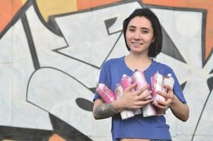 Portrait of an emotional young girl with black hair and piercings. Photo of a girl with aerosol paint cans in hands on a graffiti wall background. The concept of street art and use of aerosol paints