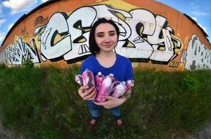 Portrait of an emotional young girl with black hair and piercings. A wide-angle photo of a girl with aerosol paint cans in the hands on a graffiti wall background. A modern portrait of a fisheye lens