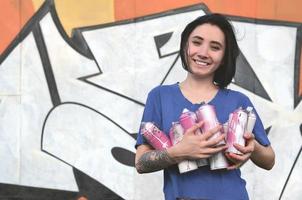 Portrait of an emotional young girl with black hair and piercings. Photo of a girl with aerosol paint cans in hands on a graffiti wall background. The concept of street art and use of aerosol paints