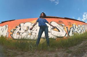 Portrait of an emotional young girl with black hair and piercings. A wide-angle photo of a girl with aerosol paint cans in the hands on a graffiti wall background. A modern portrait of a fisheye lens