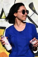 Portrait of an emotional young girl with black hair and piercings. Photo of a girl with aerosol paint cans in hands on a graffiti wall background. The concept of street art and use of aerosol paints