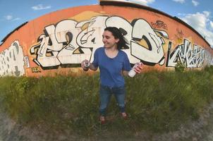 Portrait of an emotional young girl with black hair and piercings. A wide-angle photo of a girl with aerosol paint cans in the hands on a graffiti wall background. A modern portrait of a fisheye lens