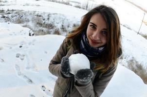 A young and joyful Caucasian girl in a brown coat holds a snowball in front of a horizon line between the sky and a frozen lake in winter. Fisheye Photo