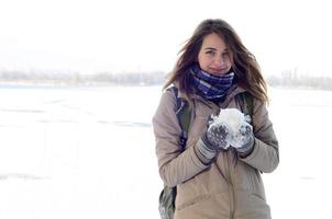 una joven y alegre chica caucásica con un abrigo marrón sostiene una bola de nieve en el fondo de una línea de horizonte entre el cielo y un lago congelado en invierno foto