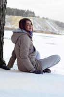 una joven caucásica con un abrigo marrón está sentada cerca de un acantilado en el fondo de una línea de horizonte entre el cielo y un lago congelado en invierno foto