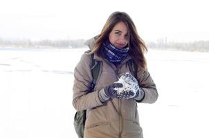 A young and joyful Caucasian girl in a brown coat holds a snowball in the background of a horizon line between the sky and a frozen lake in winter photo