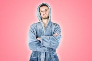 Portrait of young caucasian bearded man in blue bathrobe with crossed hands isolated on pink background photo