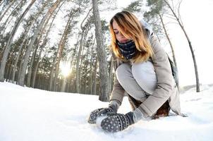 A young and joyful Caucasian girl in a brown coat sculpts a snowball in a snow-covered forest in winter. Games with snow in the open air. Fisheye Photo