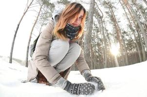 A young and joyful Caucasian girl in a brown coat sculpts a snowball in a snow-covered forest in winter. Games with snow in the open air. Fisheye Photo