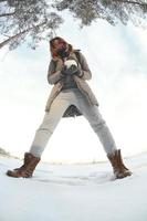 A young and joyful Caucasian girl in a brown coat holds a snowball in front of a horizon line between the sky and a frozen lake in winter. Fisheye Photo