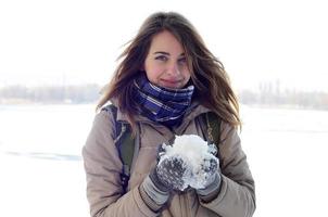 A young and joyful Caucasian girl in a brown coat holds a snowball in the background of a horizon line between the sky and a frozen lake in winter photo