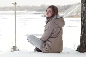 A young Caucasian girl in a brown coat is sitting near a cliff in the background of a horizon line between the sky and a frozen lake in winter time photo