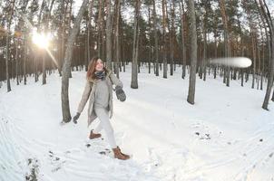 A young and joyful Caucasian girl in a brown coat is throwing a snowball in a snow-covered forest in winter. Games with snow in the open air. Fisheye Photo