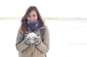 A young and joyful Caucasian girl in a brown coat holds a snowball in the background of a horizon line between the sky and a frozen lake in winter photo