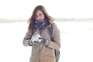 A young and joyful Caucasian girl in a brown coat holds a snowball in the background of a horizon line between the sky and a frozen lake in winter photo