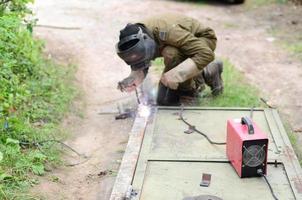 soldador anciano con uniforme marrón, máscara de soldadura y pieles de soldador, puerta de metal soldado con máquina de soldadura por arco al aire libre foto