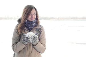 A young and joyful Caucasian girl in a brown coat holds a snowball in the background of a horizon line between the sky and a frozen lake in winter photo