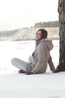 A young Caucasian girl in a brown coat is sitting near a cliff in the background of a horizon line between the sky and a frozen lake in winter time photo