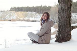 A young Caucasian girl in a brown coat is sitting near a cliff in the background of a horizon line between the sky and a frozen lake in winter time photo