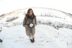 una joven y alegre chica caucásica con un abrigo marrón sostiene una bola de nieve frente a una línea de horizonte entre el cielo y un lago congelado en invierno. foto de ojo de pez