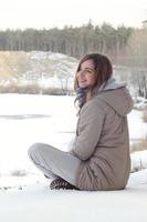 A young Caucasian girl in a brown coat is sitting near a cliff in the background of a horizon line between the sky and a frozen lake in winter time photo