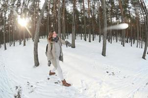 A young and joyful Caucasian girl in a brown coat is throwing a snowball in a snow-covered forest in winter. Games with snow in the open air. Fisheye Photo