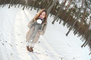 A young and joyful Caucasian girl in a brown coat holds a snowball in a snow-covered forest in winter. Fisheye Photo