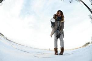 una joven y alegre chica caucásica con un abrigo marrón sostiene una bola de nieve frente a una línea de horizonte entre el cielo y un lago congelado en invierno. foto de ojo de pez