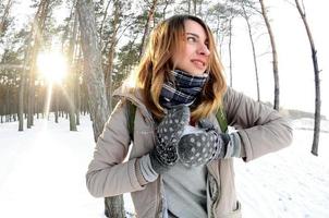 A young and joyful Caucasian girl in a brown coat sculpts a snowball in a snow-covered forest in winter. Games with snow in the open air. Fisheye Photo