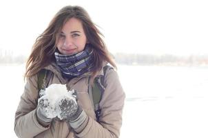 A young and joyful Caucasian girl in a brown coat holds a snowball in the background of a horizon line between the sky and a frozen lake in winter photo