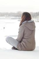 A young Caucasian girl in a brown coat staring into the distance on the horizon line between the sky and the frozen lake in winter photo