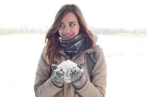 A young and joyful Caucasian girl in a brown coat holds a snowball in the background of a horizon line between the sky and a frozen lake in winter photo