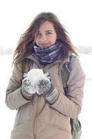 A young and joyful Caucasian girl in a brown coat holds a snowball in the background of a horizon line between the sky and a frozen lake in winter photo