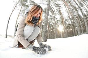 A young and joyful Caucasian girl in a brown coat sculpts a snowball in a snow-covered forest in winter. Games with snow in the open air. Fisheye Photo