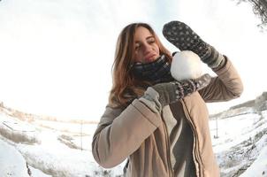 A young and joyful Caucasian girl in a brown coat holds a snowball in front of a horizon line between the sky and a frozen lake in winter. Fisheye Photo