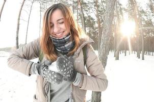 A young and joyful Caucasian girl in a brown coat sculpts a snowball in a snow-covered forest in winter. Games with snow in the open air. Fisheye Photo