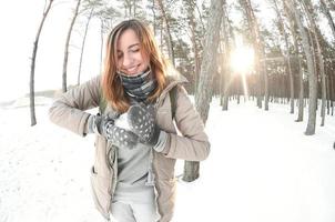 una joven y alegre chica caucásica con un abrigo marrón esculpe una bola de nieve en un bosque cubierto de nieve en invierno. juegos con nieve al aire libre. foto de ojo de pez