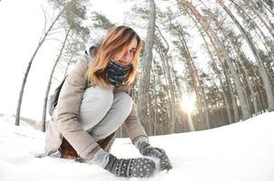 una joven y alegre chica caucásica con un abrigo marrón esculpe una bola de nieve en un bosque cubierto de nieve en invierno. juegos con nieve al aire libre. foto de ojo de pez