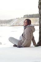 A young Caucasian girl in a brown coat is sitting near a cliff in the background of a horizon line between the sky and a frozen lake in winter time photo