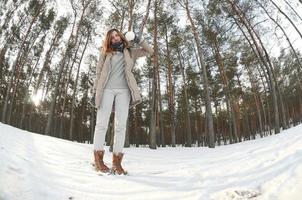 A young and joyful Caucasian girl in a brown coat holds a snowball in a snow-covered forest in winter. Fisheye Photo