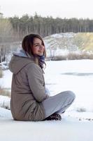 A young Caucasian girl in a brown coat is sitting near a cliff in the background of a horizon line between the sky and a frozen lake in winter time photo