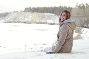 A young Caucasian girl in a brown coat is sitting near a cliff in the background of a horizon line between the sky and a frozen lake in winter time photo